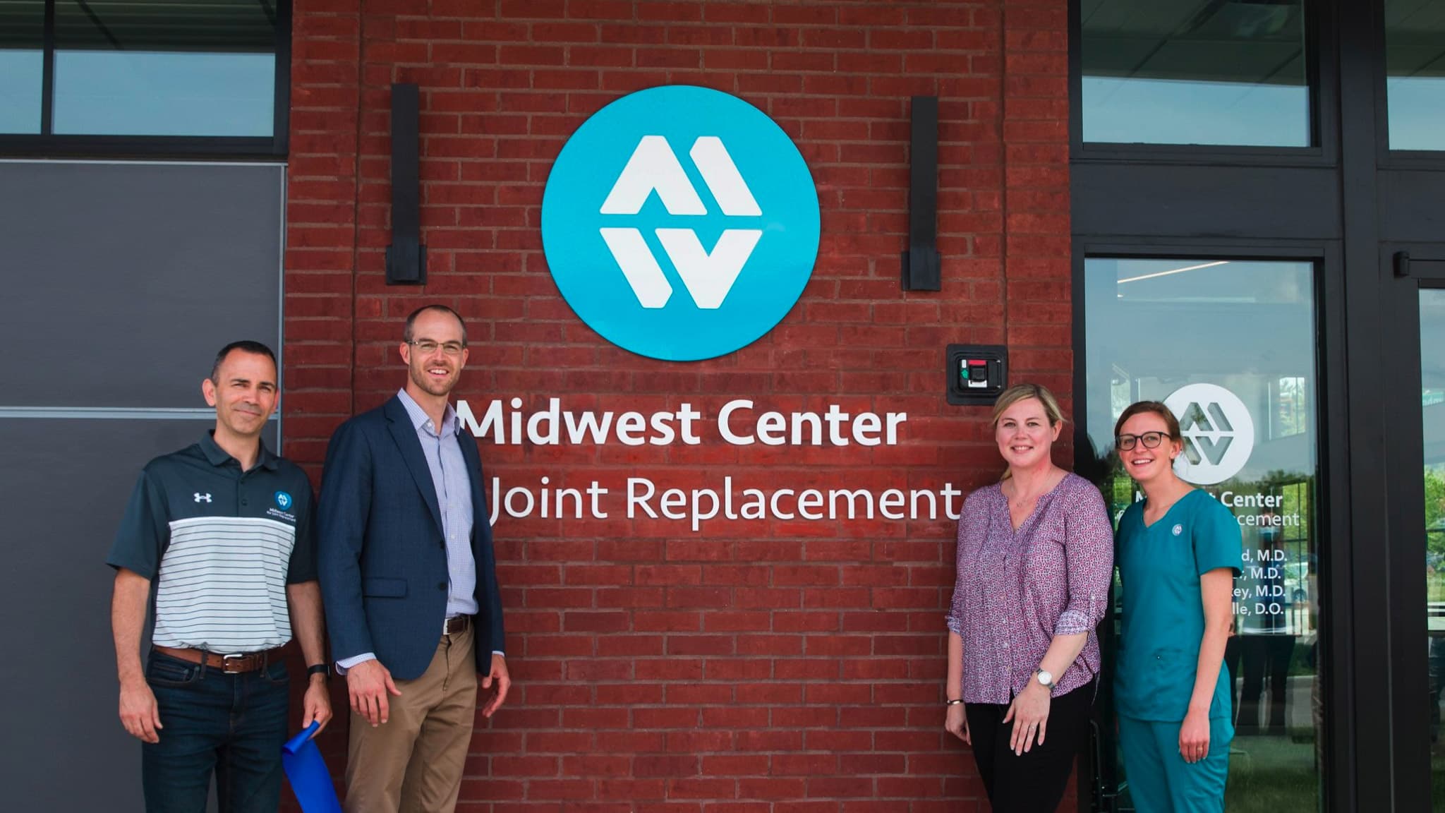 Staff from the Midwest Center for Joint Replacement stand in front of a company sign on the front of a MCJR location